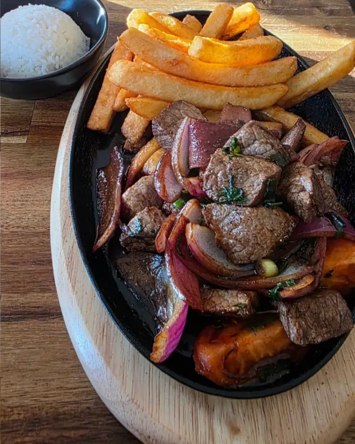 Lomo saltado sizzling on an iron plate with chips, red onion and tomato